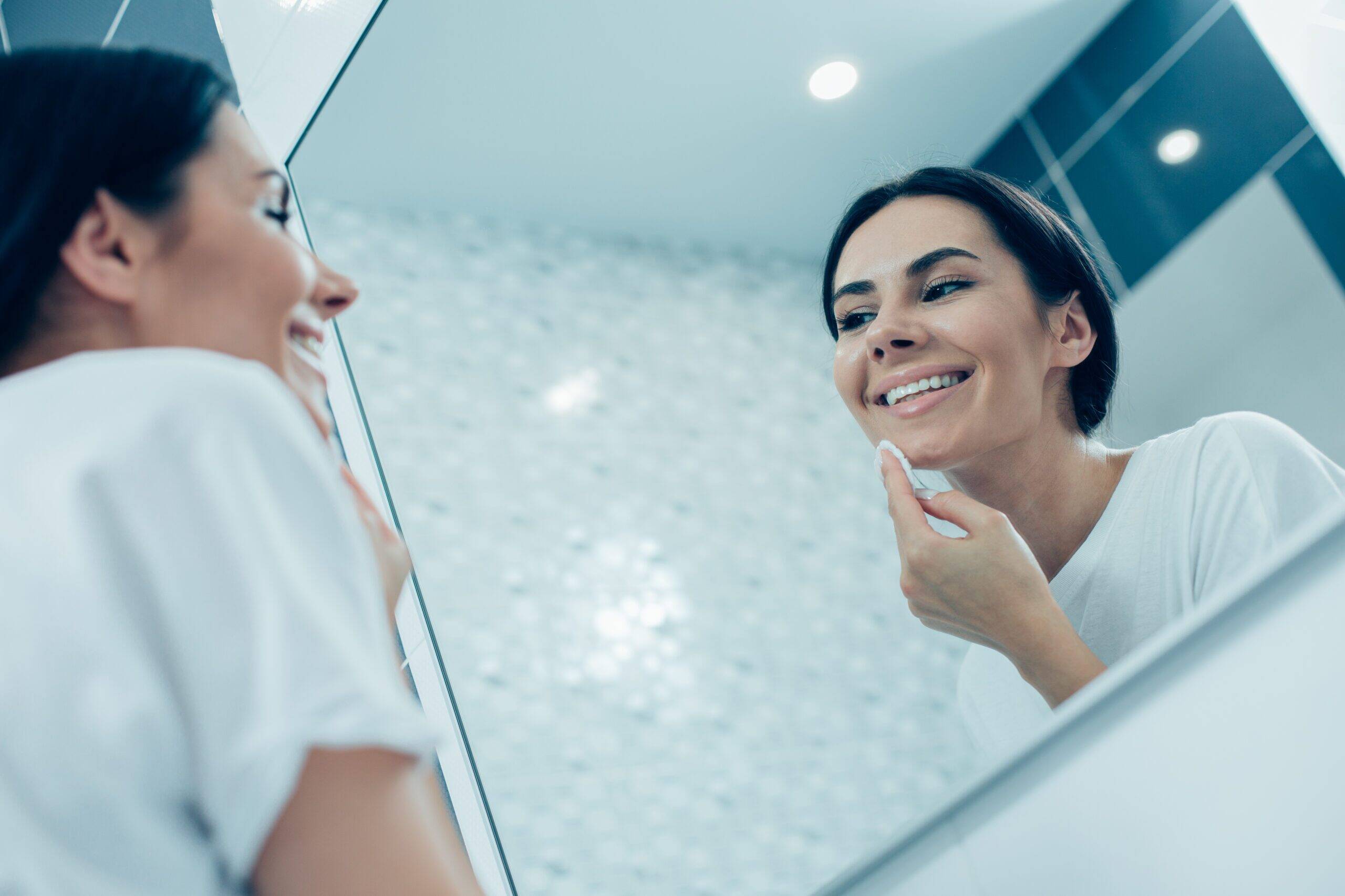 Contented young lady smiling and looking in the mirror and cleaning her face with a cotton pad