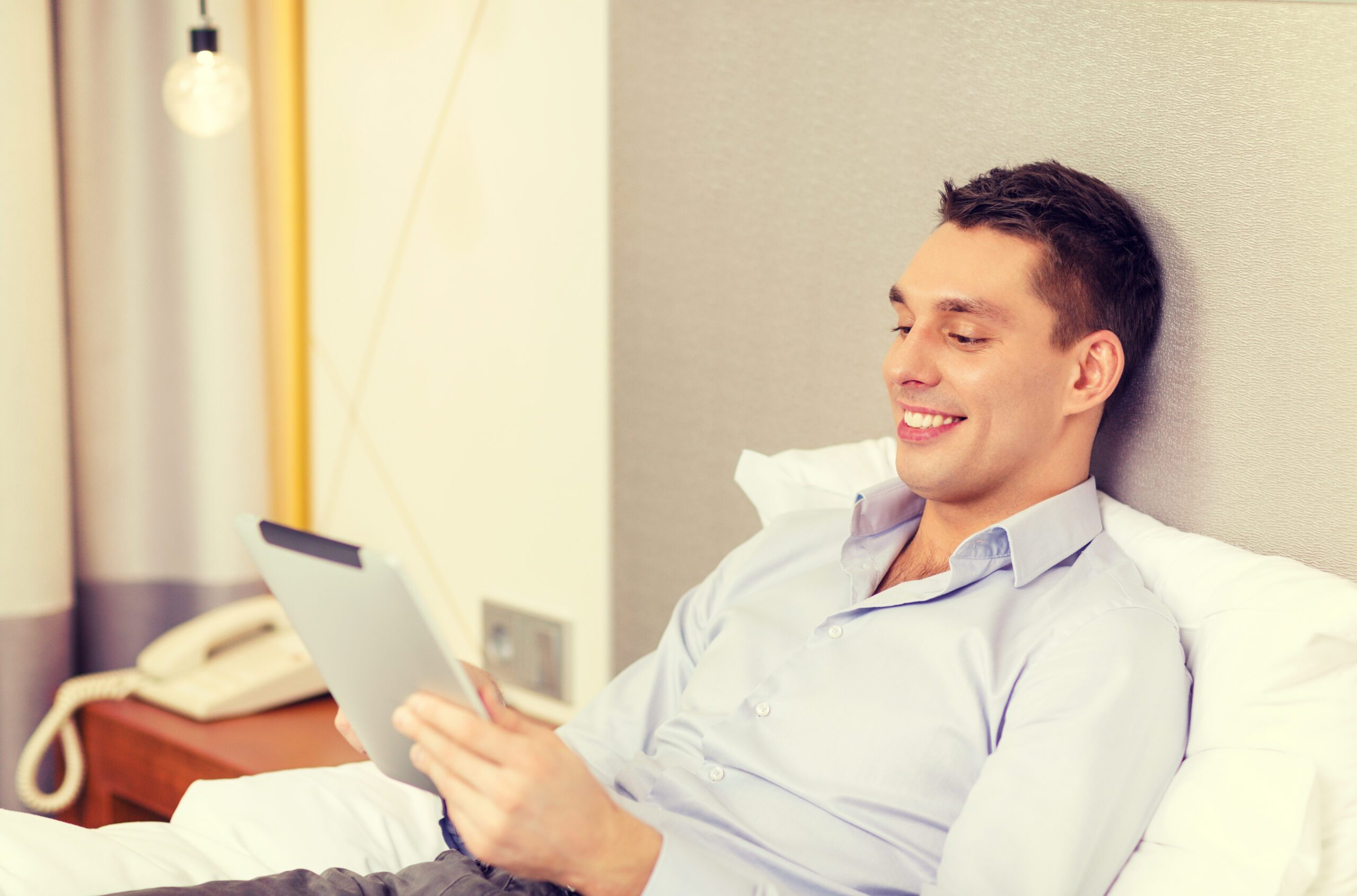 business, technology, internet and hotel concept - happy businessman with tablet pc computer lying in hotel in bed