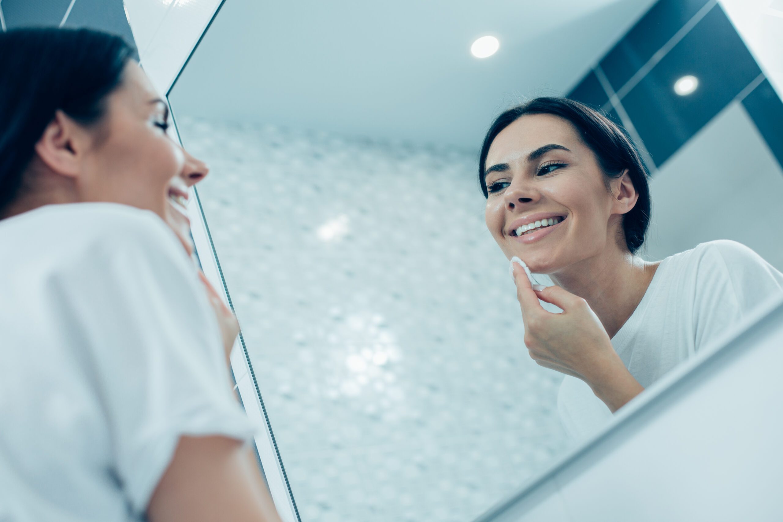 Contented young lady smiling and looking in the mirror and cleaning her face with a cotton pad