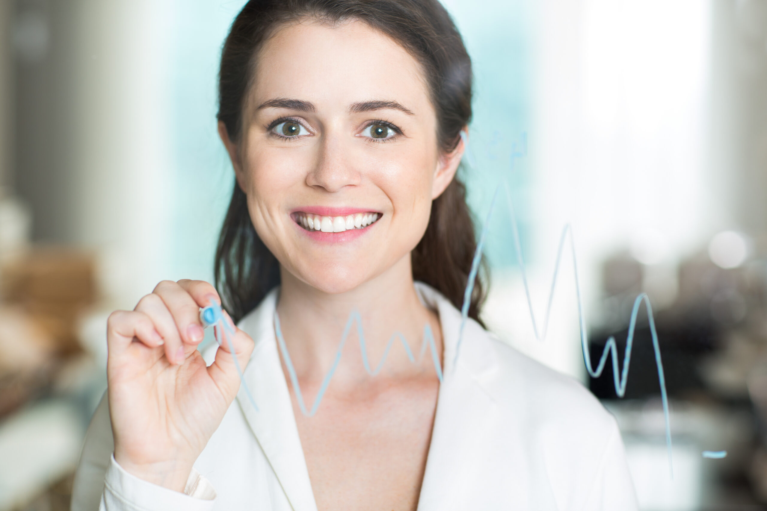 Closeup portrait of smiling at camera pretty adult businesswoman drawing graph on transparent glass and standing behind it with blurry office interior in background