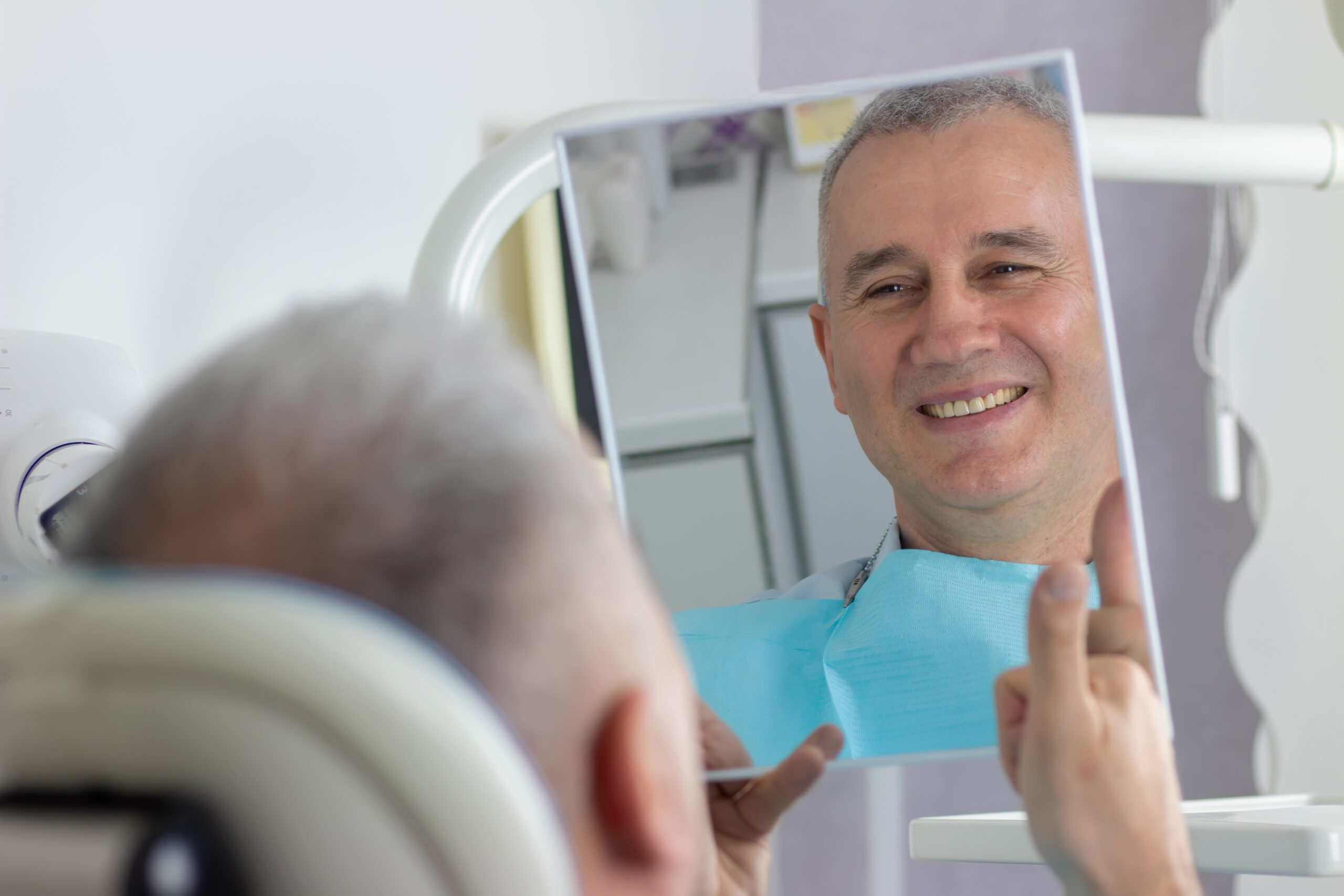A middle-aged man looks in the mirror with a healthy smile in the dentist's office.
