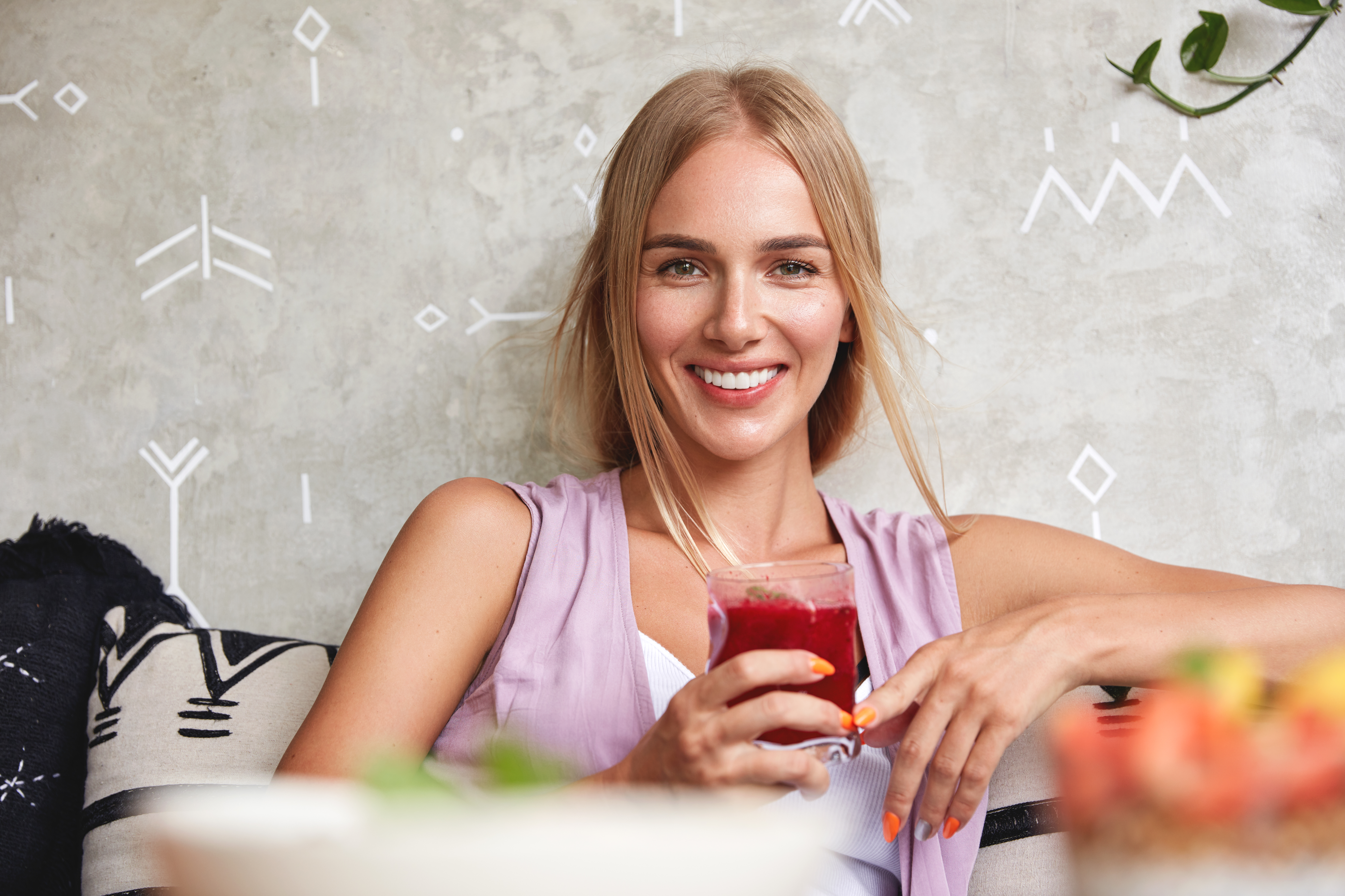 Positive beautiful young female model feels relaxed while sits on comfortable sofa in cafe, enjoys fresh strawberry smoothie, recreats during summer holidays. People, rest and lifestyle concept