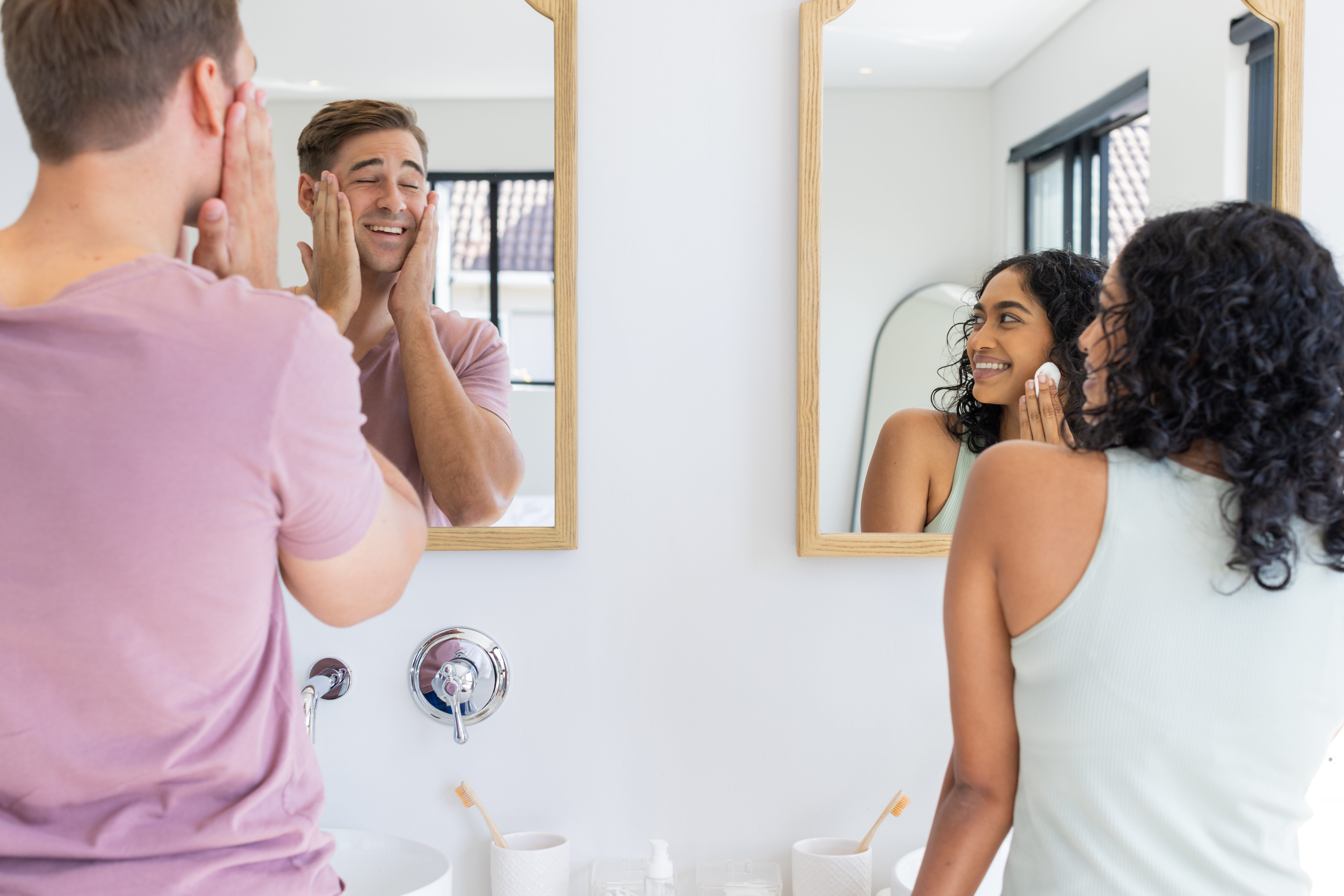 Smiling couple in bathroom, applying skincare products while looking in mirror. Beauty, routine, morning, grooming, cosmetics, wellness